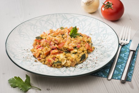 Dinkelrisotto - Auf einem runden grün-gemusterten Teller ist eine Portion Dinkelrissoto angerichtet. Rechts neben dem Teller liegen Messer und Gabel auf einer Serviette. Im Hintergrund liegt eine Tomate und eine Zwiebel. Im Vordergrund ein Blatt Petersilie. (Foto: VrK/Alexander Stiegler - Nicht zur freien Verwendung)