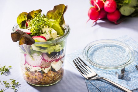 Dinkelsalat im Glas - Der bunte Schichtsalat ist in einem Gals angerichtet. Die Schichten Dinkel, Schafkäse, Radieschen, Salat und darauf Kresse sind gut zu erkennen. Neben dem Glas liegt der Deckel. Im Hintergrund liegt ein Bund Radieschen. Ebenso neben dem Glas liegt eine Gabel. (Foto: VrK/Alexander Stiegler - Nicht zur freien Verwendung)