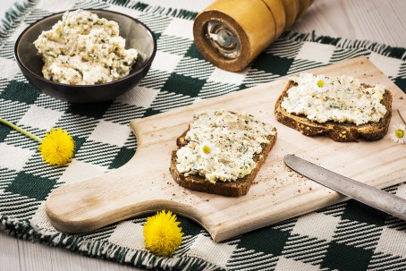 Grünkernaufstrich - Auf einem schwarz-weiß karriertem Tuch steht ein Holzbrett mit Haltegriff. Auf dem Brett liegen 2 Scheiben Schwarzbrot, die mit einem hellen Aufstrich bedeckt sind. Als Deko liegen Gänseblümchen und Löwenzahnblüten verteilt. Im Hintergrund steht ein blaues Schüsserl mit dem restlichen Aufstrich darin. (Foto: VrK/Alexander Stiegler - Nicht zur freien Verwendung)