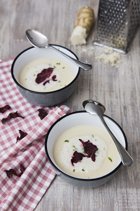 Krenschaumsuppe mit Rote-Rüben-Chips - Auf einem grauen Tisch sstehen zwei Schalen mit scharzem Rand, daneben liegt ein rot/weiß kariertes Tuch; in den Schalen befindet sich ein weiße, aufgeschäumte Suppe, garniert mit Roten-Rüben-chips; daneben noch ein Stück Kren mit einer Reibe und zwei Löffel (Foto: Barbara Majcan - Nicht zur freien Verwendung)