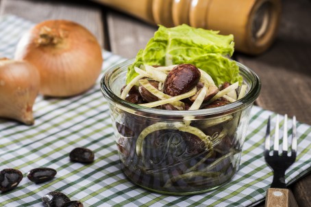 Käferbohnensalat - Auf einem grün/weiß karierten Tischtuch steht eine Glasschüssel mit dunklen rot/braunen Käferbohnen, garniert mit weißen Zwiebeln, grünen Salat und Kürbiskernöl; daneben liegen ungeschälte gelbe Zwiebel und einige einzelne Käferbohnen (Foto: VrK/Alexander Stiegler - Nicht zur freien Verwendung)