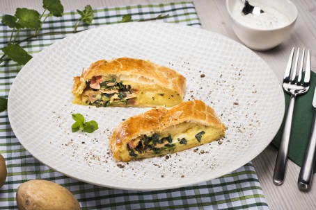 Pikanter Bärlauchstrudel mit Schinken und Erdäpfel - Auf einem weißen Teller liegen zwei Stück goldgelb gebackener Strudelt mit einer grün/rosa Füllung, bestehend aus Bärlauch und Schinken. Das ganze ist mit etwas Kräutersalz bestreut. Daneben liegt ein Besteck, eine Schale mit einem weißen Dip und eines grün/weiße Serviette. (Foto: VrK/Alexander Stiegler - Nicht zur freien Verwendung)