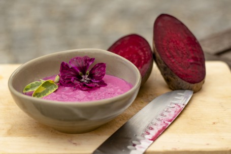 Rote Rüben Tsatsiki mit Kren - Auf einem Holzbrett ist in einer graubraunen Schüssel das lilafarbige Rote Rüben Tsatsiki angerichtet. Eine lilafarbige Blüte und 2 grüne blätter sind als Deko darauf platziert. Im Hintergrund ist eine Rote Rübe die in der Mitte aufgeschnitten ist positioniert. Rechts neben der Schüssel liegt ein Messer. (Foto: Tobias Schneider-Lenz - Nicht zur freien Verwendung)