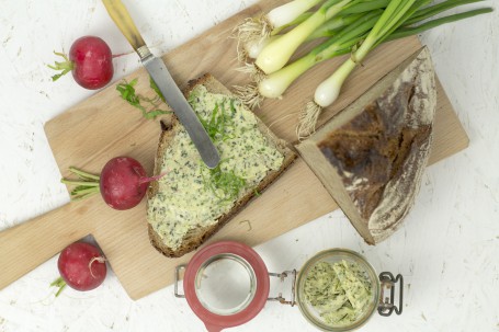 Bunte Kräuterbutter - Auf einem Holzbrett mit Stiehl liegt eine Schnitte Brot, die mit der Kräuterbutter bestrichen ist. Die grünen fein gehackten Kräuter sind deutlich zu erkennen. Auf das Brot sind noch grob gehackte Kräuter gestreut. Auf dem Teller liegen noch einige Jungzwiebeln, ein Radieschen und ein Brotscherz. Neben dem Teller befinden sich noch 2 Radieschen und ein kleines Rex-Glas mit Kräuterbutter darin. (Foto: Tobias Schneider-Lenz - Nicht zur freien Verwendung)