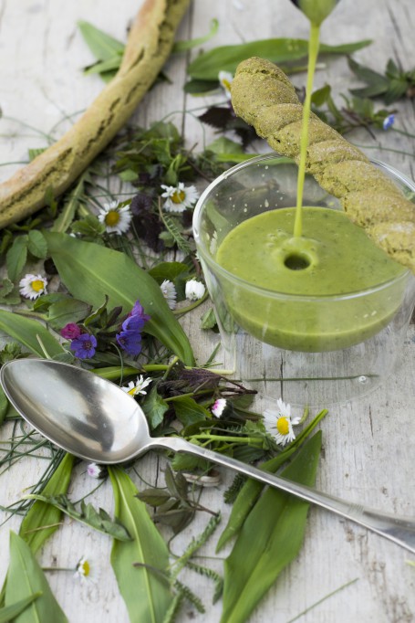 Wildkräutersupperl - In einem Glasschüsserl ist die grüne Cremesuppe angerichtet. Am Bild wird noch Suppe eingegossen. Schräg über dem Glasschüsserl liegt ein Grissinistangerl. Rund um das Gefäß sind Wildkräuter und Blüten verstreut. Ein silberner Esslöffel liegt im Vordergrund. (Foto: Tobias Schneider-Lenz - Nicht zur freien Verwendung)