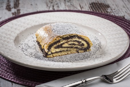 Mohnstrudel aus Erdäpfelteig - Auf einem lila Platzset steht ein weißes Teller; darauf liegt ein aufgeschnittenes Stück eines goldgelb gebackenen Mohnstrudels, bestreut mit Zucker/Mohngemisch; daneben liegt eine weiße Serviette und eine Gabel (Foto: VrK/Alexander Stiegler - Nicht zur freien Verwendung)