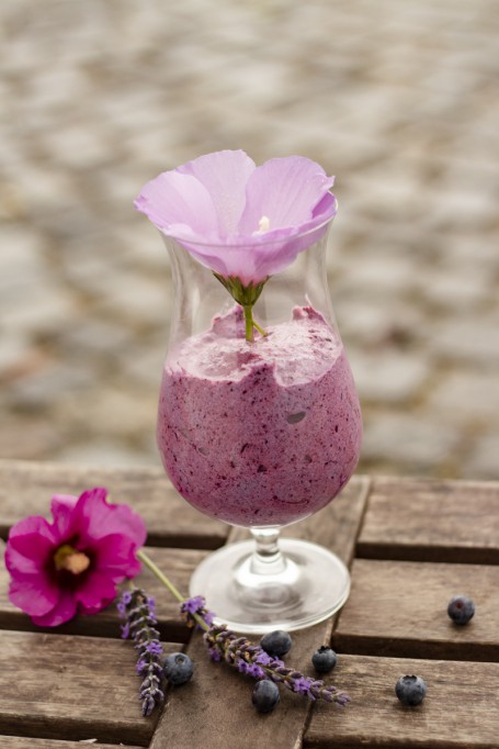 Luftiger Beeren-Hexenschaum - Auf einem Holzuntergrund steht in einem Cocktailglas serviert das lilafarben Dessert. Garniert ist das Dessert mit einer helllila Blüte. Vor dem Glas liegen einige Heidelbeeren, Lavendelblüten und eine dunkellila-farbige Blüte. (Foto: Tobias Schneider-Lenz - Nicht zur freien Verwendung)