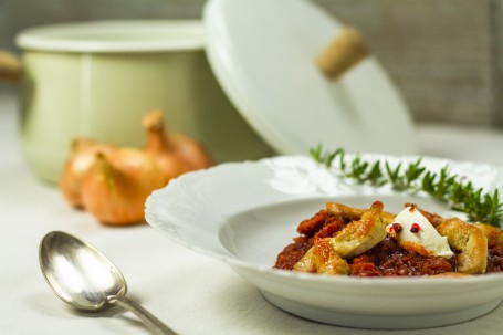 Hendl-Kürbis-Gulasch - In einem weißen Suppenteller ist das rötliche Hendl-Gulasch angerichtet. Die Hendl-Stücke sind hellbraun angeröstet und auf dem Gulasch platziert. Ein Tupen Sauerrahm ist als Dekoration am Gulasch angerichtet. Ein Kräuterzweig befindet sich am Tellerrand im Hintergrund. Hinter dem Teller sind verschwommen ein grünlicher Kochtopf und 3 Stück Zwiebel zu sehen. Links neben dem Suppenteller liegt ein Suppenlöffel bereit. (Foto: Tobias Schneider-Lenz - Nicht zur freien Verwendung)