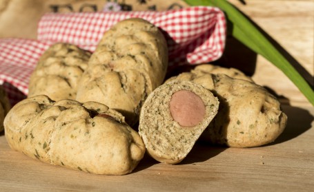 Würstel im Dinkel-Kräuterteig - Auf dem Bild sind vier Stück der Würstel im Brotteig zu sehen. Ein halbes Weckerl liegt im Vordergrund. In der Mitte ist das Würstchen zu sehen. Im Hintergrund des Bildes liegt ein rot-weiß-karriertes Tuch. (Foto: Romana Schneider-Lenz - Nicht zur freien Verwendung)