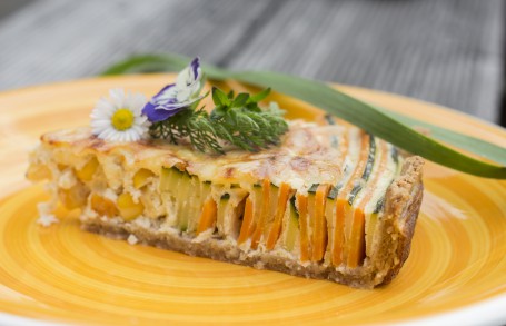 Einkorn-Gemüse-Quiche - Auf einem gelben Teller ist ein Stück der Gemüse Quiche angerichtet. Die abwechselnden Gemüseschichten sind deutlich zu erkennen und sehen sehr bunt aus. Der Überguss ist auf der Oberfläche leicht bräunlich. Auf dem Stück liegt ein Gänseblümchen und eine lila Blüte als Deko. Auch grüne Kräuterzweige sind dekorativ darauf platziert. (Foto: Moritz Lenz - Nicht zur freien Verwendung)