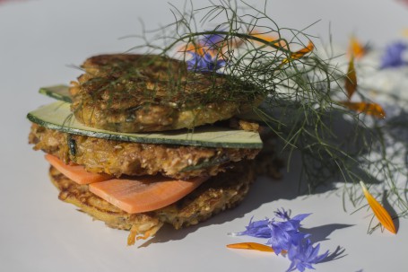 Pikante Getreidelaibchen - Auf dem Bild sind drei Getreidelaibchen aufeinander gestapelt. Dazwischen sind Zucchini und Karottenstreifen gelegt.Über den Laibchen liegt eine Kräuterzweig. Rechts vor den Laibchen liegen einige bunte Blüten verstreut. (Foto: Schneider-Lenz - Nicht zur freien Verwendung)