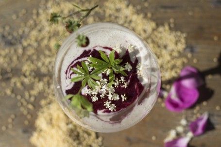 Hirse-Topfen-Joghurt-Creme - Auf einer Holzfläche steht ein Weinglas in dem das Dessert angerichtet ist. Es ist gräulich. An der Oberfläche ist das Fruchtmus zu erkennen. Über das Fruchtmus sind einzelne Hollunderblüten gestreut. Auch auf der Holzfläche sind einzelne Hollunderblüten und auch andere bunte Blüten verstreut. (Foto: Schneider-Lenz - Nicht zur freien Verwendung)