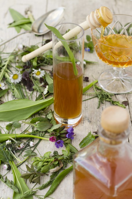 Oxymel - Am Bild liegen auf einer hellen Holzplatte diverse Blüten und Wildkräuter. Im Vordergrund steht eine eckige Flasche in der sich das fertige Oxymel befindet. In der Mitte des Bildes steht ein hohes Glas das zur Hälfte damit gefüllt ist. Ein grüner Halm befindet sich im Glas. Im hinteren Teil des Bildes steht eine Sektschale an der ein Honigportionierer angelehnt ist. Der Honig darauf tropft in das Glas. (Foto: Tobias Schneider-Lenz - Nicht zur freien Verwendung)