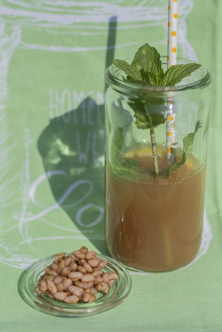 Barley Water - Ein hohes Glas ist zur Hälfte mit dem bräunlichen Gerstenwasser befüllt. Darin befindet sich ein Kräuterzweig und ein weißer Strohhalm mit gelben Punkten. Vor dem Glas liegt der Deckel auf dem sich Gerstenkörner befinden. (Foto: Tobias Schneider-Lenz - Nicht zur freien Verwendung)