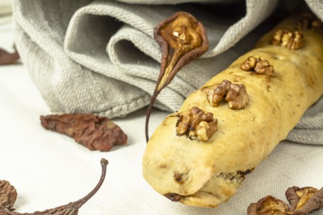 Früchtebrot im Mantel - Auf dem Bild ist ein längliches Früchtebrot in einem dünnen Teigmantel zu sehen. Auf dem Brot sind vier Nüsse zu sehen, die mitgebacken wurden. Verwinzelt liegen getrocknete Birnenhälften um das Brot herum. Das Brot ist zur hälfte in ein graues Tuch gewickelt, das im Hintergrund zu erkennen ist. (Foto: Tobias Schneider-Lenz - Nicht zur freien Verwendung)