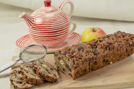 Apfel-Nuss-Brot - Auf einem Holzbrett liegt das längliche Apfel-Nuss-Brot. Die Apfel,- und Nussstückchen sind darin gut zu erkennen. Drei Scheiben des Brotes sind bereits abgeschnitten und liegen schräg leicht überlappend am Brett. Hinter den Scheiben liegt ein kleines Sieb. Hinter dem Brett steht eine rot-weiß-gestreifte Teekanne auf einer dazu passenden Untertasse. Ebenso im hintergrund liegt ein Apfel. (Foto: Tobias Schneider-Lenz - Nicht zur freien Verwendung)