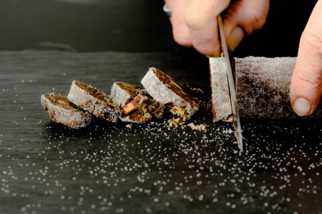 Siaße Blunzn - Auf einer schwarzen Fläche liegen vier Scheiben der dünnen Rolle. Ein fünftes Stück wird gerade heruntergeschnitten. Der Rest der Rolle ist auch am Bild zu sehen. Die Rolle ist von weißem Zucker leicht überzogen. Im Vordergrund ist Zucker leicht auf der Fläche verstreut. (Foto: Tobias Schneider-Lenz - Nicht zur freien Verwendung)