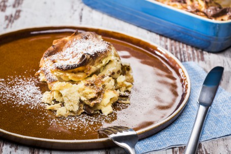 Topfenpalatschinken überbacken - Auf blau/braunen Hintergrund steht ein brauner Teller, auf dem sich eine goldgelb gebackenen, in Form eines Türmchens angerichtete, Palatschinke befindet. Garniert mit Staubzucker, daneben steht eine blaue Auflaufform und ein Besteck. (Foto: VrK/Alexander Stiegler - Nicht zur freien Verwendung)