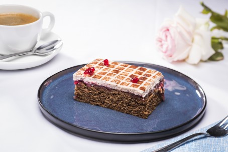 Käferbohnen-Preiselbeerschnitte - Auf einem runden blauen Teller ist ein rechteckiges Stück der Schnitte platziert. Auf dem dunklen Boden ist eine dünne Schicht der weißen Creme aufgetragen. Auf der Creme ist mit Kakaopulver ein Muster aus lauter kleinen Quadraten gestreut. Drei Preiselbeeren leigen auf dem Kuchenstück. Im Hintergrund liegt eine weiße leicht rosa schimmernde Rose und eine Tasse Kaffee mit Untertasse und Löffel sind dort platziert. (Foto: VrK/Alexander Stiegler - Nicht zur freien Verwendung)