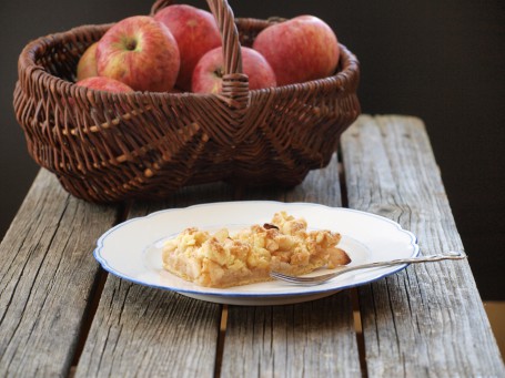 Apfel-Streuselkuchen - Auf einer Holzfläche steht ein weißer Teller mit blauem Rahmen, auf dem sich ein Stück des Apfel-Streuselkuchen befindet. Neben dem Kuchenstück liegt eine silberne Dessertgabel. Im Hintergrund steht ein großer brauner geflochtener Korb mit großen rotbackigen Äpfeln darin. (Foto: VrK/DI Carina Laschober- Luif - Nicht zur freien Verwendung)