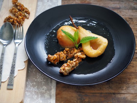 Birnendessert mit karamellisierten Walnüssen - Auf einer holzfläche steht ein runder schwarzer Teller auf dem zwei Birnenhälften angerichtet sind. Die Birnen sind mit der durchsichtigen Flüssigkeit übergossen und mit einem Minzeblatt dekoriert. Im vorderen Teil des Tellers sind einige der karamellisierten Nüsse, die in zwei Streifen gelegt sind angerichtet. Links neben dem Teller steht ein Holzbrett auf dem ein Löffel und eine Gabel zu sehen sind. Im hinteren Teil des Brettes liegen einige karamelisierte Nüsse. (Foto: VrK/DI Carina Laschober- Luif - Nicht zur freien Verwendung) Birnendessert mit karamellisierten Walnüssen - Auf einer holzfläche steht ein runder schwarzer Teller auf dem zwei Birnenhälften angerichtet sind. Die Birnen sind mit der durchsichtigen Flüssigkeit übergossen und mit einem Minzeblatt dekoriert. Im vorderen Teil des Tellers sind einige der karamellisierten Nüsse, die in zwei Streifen gelegt sind angerichtet. Links neben dem Teller steht ein Holzbrett auf dem ein Löffel und eine Gabel zu sehen sind. Im hinteren Teil des Brettes liegen einige karamelisierte Nüsse. (Foto: VrK/DI Carina Laschober- Luif - Nicht zur freien Verwendung)