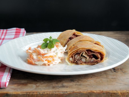 Bohnenstrudel mit Apfel-Karottensalat - Auf einer Holzfläche steht ein weißer Teller auf dem drei Stücke des Bohnenstrudels hinterinander aufgelegt sind. Die Bohnen sind im Strudel zu erkennen. Neben den Strudelstücken ist eine Portion des Apfel-Karottensalates angerichtet. Auf dem Salat liegt als Garnitur ein Minzeblatt. (Foto: VrK/DI Carina Laschober- Luif - Nicht zur freien Verwendung)