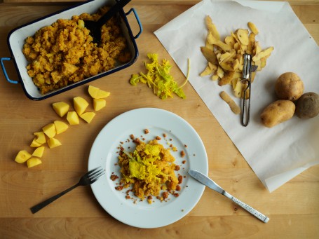 Erdäpfel- Polentasterz - Das Bild zeigt einen weißen Teller von oben, auf dem eine Portion des gelblichen Sterz angerichtet ist. Darüber liegen die Blätter der Halmrübe. Links oben steht eine blaue Auflaufform mit zwei Hänkeln, in der sich der Rest des Sterzes befindet. Rechts oben liegen auf einem Papier drei ungeschälte Erdäpfel und einige Schalen mit einem Schäler. Zwischen Auflaufform und Teller liegen einige rohe Kartoffelstücke und ein paar Blätter der Halmrübe. (Foto: VrK/DI Carina Laschober- Luif - Nicht zur freien Verwendung)