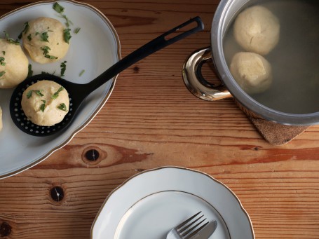 Selchfleischknödel - Auf einem Holztisch steht ein silberner Suppentopf, der zur Hälfte mit Wasser gefüllt ist. Zwei selchfleischknödel schwimmen darin. In der linken Bildhälfte steht eine weiiße Porzellanplatte auf der einige fertige Knödel liegen und mit gehackter Petersilie bestreut sind. Im Vordergrund des Bildes steht ein weißer Teller mit goldener Verziwhrung auf dem sich Messer und Gabel befinden. (Foto: VrK/DI Carina Laschober- Luif - Nicht zur freien Verwendung)