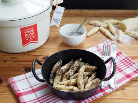 Mohnnudeln - In einer kleinen schwarzen Pfanne mit zwei Hänkeln befindet sich eine Portion Mohnnudeln. Die Pfanne steht auf einem weiß-rot karriertem Tuch. Rechts neben der Pfanne auch auf dem Tuch liegen Messer und Gabel bereit. Im Hintergrund steht ein kkeines weißees Schüsserl in dem sich das Mohn-Zucker-Gemisch befindet. Rchts neben dem Schüsserl liegen einige gedrehte Schupfnudeln und der Rest des Teiges. Liks neben dem Schüsselchen steht ein weißer Kochtopf mit roter Aufschrift. (Foto: VrK/DI Carina Laschober- Luif - Nicht zur freien Verwendung)