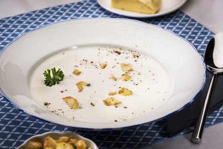 Steirerkässuppe - Auf blauem Tischset steht ein weißer Teller mit einer weißen Suppe, garniert mit Brotwürfeln und Krfäutern. Daneben liegt ein Löffel und eine Schale mit weiteren goldgelbenen Brotwürfeln. (Foto: VrK/Alexander Stiegler - Nicht zur freien Verwendung)