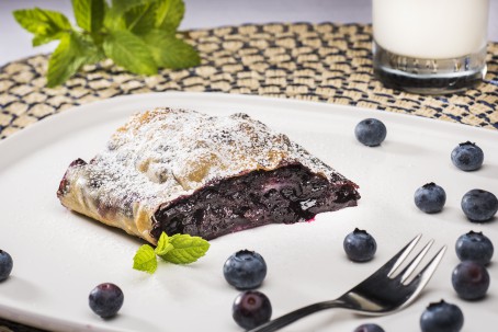 Schwarzbeerstrudel - Auf weißem Teller liegt ein goldgelb gebackenes Teil eines Strudels, mit Staubzucker garniert; die Füllung ist dunkellila. Daneben liegen einige Heidelbeeren und ein Melissenblatt. Daneben steht ein Glas Milch und ein Zweig Melisse. (Foto: VrK/Alexander Stiegler - Nicht zur freien Verwendung)