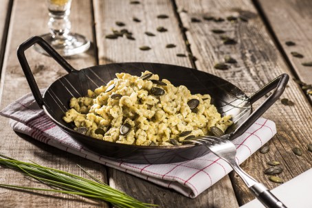 Kürbiskernspätzle - Auf einem weiß/rot karierten Geschirrtuch steht eine schwarze Pfanne mit goldgelb farbenen Spützel, garniert mit Kürbiskernen. Daneben liegt frischer grüner Schnittlauch. (Foto: VrK/Alexander Stiegler - Nicht zur freien Verwendung)