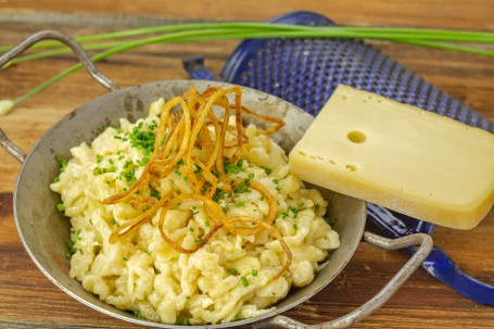 Käsespätzle - In einer Metallpfanne leigen hellgelb mit Käse überbackene Spätzle, ganiert mit gebratenen Zwiebelringen und Kräutern. Daneben liegt ein großes Stück Käse und eine blaue Reibe. (Foto: Tobias Schneider-Lenz - Nicht zur freien Verwendung)