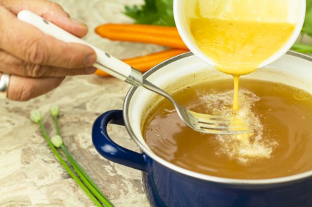 Eintropfsuppe - In einem blau/weißen Kochtopf befindet sich eine klare Flüssigkeit, in welche gerade die gelbe Eintropfmasse mit einer Gabel eingerührt wird. Daneben liegen orangefarbene Karotten. (Foto: Tobias Schneider-Lenz - Nicht zur freien Verwendung)