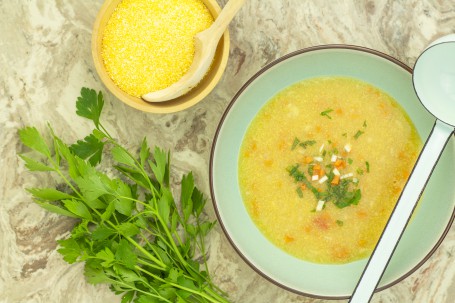 Griessuppe - In einem hellen Teller mit braunem Rand befindet sich sich die gelbe Suppe, ganiert mit Kräutern und Karotten. Daneben steht eine Holzschale mit gelb/weißem rohen Grieß und ein Bund grüne Petersilie (Foto: Tobias Schneider-Lenz - Nicht zur freien Verwendung)