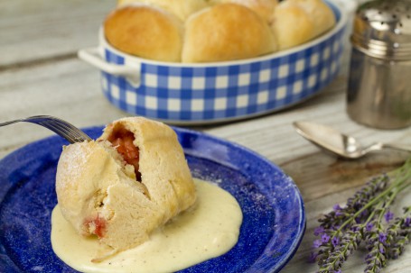 Buchteln OÖ - Auf einem blauen Teller in dem in der Mitte eine Portion Vanillesauce angerichtet ist, befindet sich eine Buchtel, die in der Mitte aufgebrochen ist. Dadurch ist die Marmeladefüllung gut zu sehen. Im Hintergrund befindet sich eine Auflaufform mit mehreren Buchteln darin. (Foto: Tobias Schneider-Lenz - Nicht zur freien Verfügung)