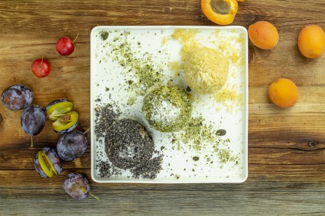 Obstknödel - Auf einem eckigen weißen Teller sind drei Knödel angerichtet. Jeder der Knödel ist mit einer anderen Zutat in den Bröseln verfeinert. Zu erkennen sind Mohn, Kürbis und nur Brösel. Rund um den Teller liegen Zwetschken, Marillen und Kirschen. (Foto: Tobias Schneider-Lenz - Nicht zur freien Verfügung)