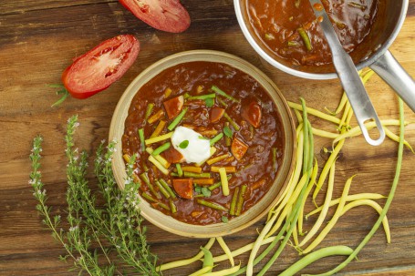 Erdäpfelgulasch - Auf einem Holztisch steht eine helle Schüssel mit braunfarbenem, cremigen Gulasch mit Stücken von Würsteln, grünen Fisolen und mit einem Klecks Sauerrahm garniert. Daneben liegen grüne Bohnen, Kräuter und rote Tomatenhälften. (Foto: Tobias Schneider-Lenz - Nicht zur freien Verwendung)