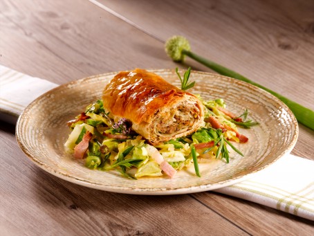 Knuspriger Leberstrudel & Leberstrudel in der Suppe - Auf einem braunen Teller liegt auf einem grünen Salatbett mit Speck ein Stück knusprig goldgelb gebackener Strudel. (Foto: VrK/Dall - Nicht zur freien Verwendung)