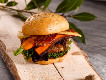 Leberburger - Auf einem Holzbrett liegt ein braun gebackenes Burgerbrötchen, gefüllt mit grünem Salat, braun gebratenem Burgerlaibchen, orangefarbenen Karotten, und knusprig gebratenem Speck. (Foto: VrK/Dall - Nicht zur freien Verwendung)