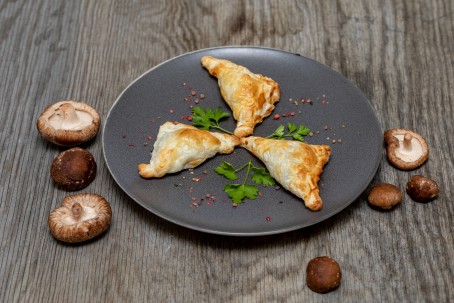 Blätterteigtaschen mit Pilzen - Auf einem dunkelgrauen Teller liegen 3 Stück Blätteteigtaschen. Dazwischen liegt jeweils ein Blatt Petersilie. Rund um den Teller sind Pilze gestreut. (Foto: VrK/Achim Mandler Photography - Nicht zur freien Verfügung)