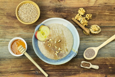 Habermus - Auf einer Holzplatte steht eine blaue runde Schüssel mit dem bräunlichen Getreidebrei darin. Der Brei ist mit etwas Zimt und Walnüssen bestreut. Am oberen Rand der Schüssel liegt eine Apfelscheibe. Rund um die Schüssel sind die Zutaten zu sehen. Honig, Nüsse, Zimtstangen und Getreideflocken sind in kleinen Gefäßen rund um die Schüssel platziert. (Foto: Tobias Schneider-Lenz - Nicht zur freien Verwendung)