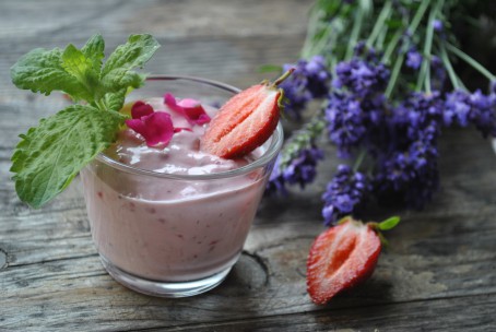 Beerige Topfencreme - Auf einer Holzplatte steht ein kleines Glas in der sich die hellrosa Topfencreme befindet. In der Creme sind kleine rote Erdbeerstückchen zu sehen. Auf der Creme ist eine halbe Erdbeere zwei rosarote Rosenblätter und drei Minzeblätter als Deko platziert. Rechts neben dem Glas liegt eine halbe Erdbeere und im Hintergrund ein Büschel Lavendel der lila blüht. (Foto: Romana Schneider-Lenz - Nicht zur freien Verwendung)