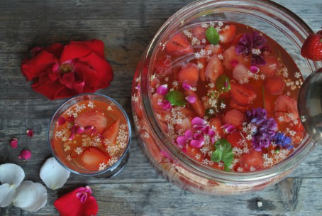 Kinderbowle - In einem großen runden Gefäß ist die Bowle angerichtet. Als Früchte wurden hier in erster Linie Erdbeeren verwendet. Die Erdbeerstückchen sind deutlich zu sehen. Es sind auch viele bunte Blüten in allen Farben und grüne Minzeblätter in der Bowle zu erkennen. Links neben dem Gefäß steht ein Glas in dem eine Portion Bowle angerichtet ist. Hinter dem Glas liegt eine große rote Blüte. (Foto: Romana Schneider-Lenz - Nicht zur freien Verwendung)