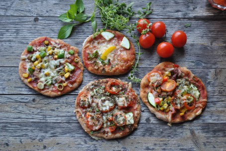 Lustiges Pizzagesicht - Auf dem Bild liegen vier verschieden belegte Pizzen. Auf einer Pizza ist ein Gesicht aus zwei Zucchinstücken als Augen, ein länglich geschnittener gelber Paprika als Nase und einige grüne Kräuter die als Mund in einem Halbkreis aufgelegt sind zu erkennen. Im Hintergrund liegen noch einige rote Tomaten und grüne Kräuter. (Foto: Romana Schneider-Lenz - Nicht zur freien Verwendung)