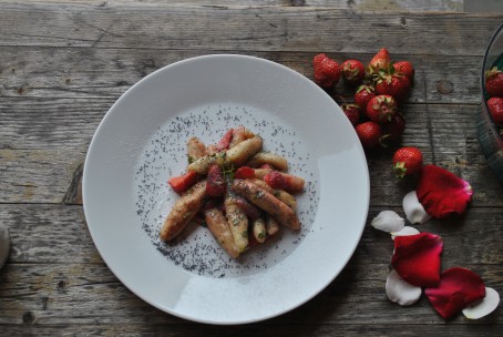 Mohnnudeln aus Erdäpfelteig - Auf dem Bild ist ein weißer runder Teller der auf einer Holzplatte steht zu sehen. Rechts neben dem Teller liegen einige Erdbeeren und weiße und rote Blütenblätter. Auf dem Teller sind einige längliche Schupfnudeln mit Mohn und Erdbeerstückchen angerichtet. Als Garnitur liegt obenauf ein grünes Minzeblatt. (Foto: Romana Schneider-Lenz - Nicht zur freien Verwendung)