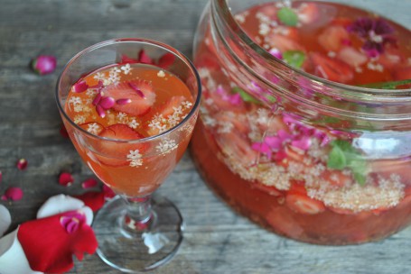 Mostbowle - Auf einer Holzfläche steht ein Stiehlglas mit der leicht rötlichen Mostbowle darin. In der Bowle schwimmen Erdbeerstücke, Holunderblüten und Rosenblüten. Neben dem Glas steht ein großes glasgefäß mit der restlichen Bowle darin. (Foto: Romana Schneider-Lenz - Nicht zur freien Verwendung)