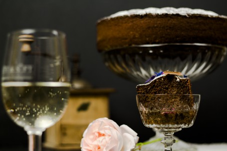 Mostschober - Rechts im Vordergrund befindet sich ein Kuchenstück in einer durchsichtigen Sektschale, die halb mit Most befüllt ist. Im Hintergrund befindet sich ein Kranzkuchen auf einem durchsichtigen hohen Teller der mit Zucker bestreut ist. Links im Vordergrund sieht man leicht verschwommen ein halb volles Mostglas stehen. In der Mitte im Vordergrund liegt eine rosarote Rose. (Foto: Tobias Schneider-Lenz - Nicht zur freien Verfügung)