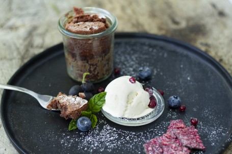 Schokokuchen - Man sieht einen schwarzen runden Teller. Darauf steht im hinteren linken Drittel ein kleines Rex-Glas in dem sich der Schokokuchen befindet. Davor liegt eine Gabel mit eiem Stück Schokokuchen darauf. Neben der Gabel liegt dekorativ ein Minzeblatt und einige Heidelbeeren. Neben der Gabel liegt der Deckel des Rex-Galses, darauf befindet sich eine Kugel Vanilleeis mit einigen Beeren dekoriert. Alles ist mit Staubzucker fein bezuckert. (Foto: Tobias Schneider-Lenz - Nicht zur freien Verfügung)