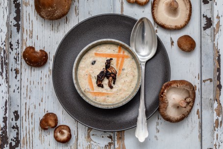 Erdäpfelsuppe mit Shiitakepilzen - Ein schwarzer Teller steht auf einem weißem Altholz Hintergrund. Rund um den Teller herum liegen einzelne Shiitakepilze. Auf dem Teller steht eine kleine Suppenschüssel in der sich die grob pürrierte Erdäpfelsuppe befindet. Die Suppe ist garniert mit grobenKarottenstiften und angebratenen Pilzen. Neben der Suppenschüssel liegt ein silberner Suppenlöffel. (Foto: VrK/Achim Mandler Photography - Nicht zur freien Verwendung)